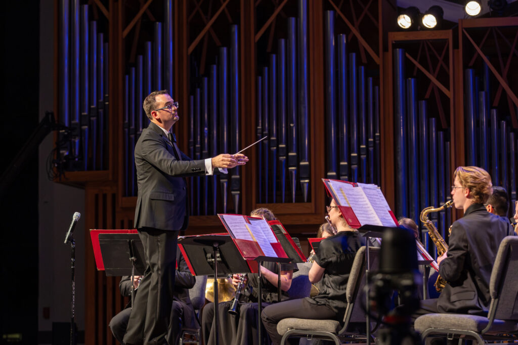 A conductor stands in front of musicians playing instruments on stage, showcasing the collaborative spirit at the heart of music education. The musicians sit with sheet music, and large organ pipes are visible in the background. Stage lights illuminate the scene.