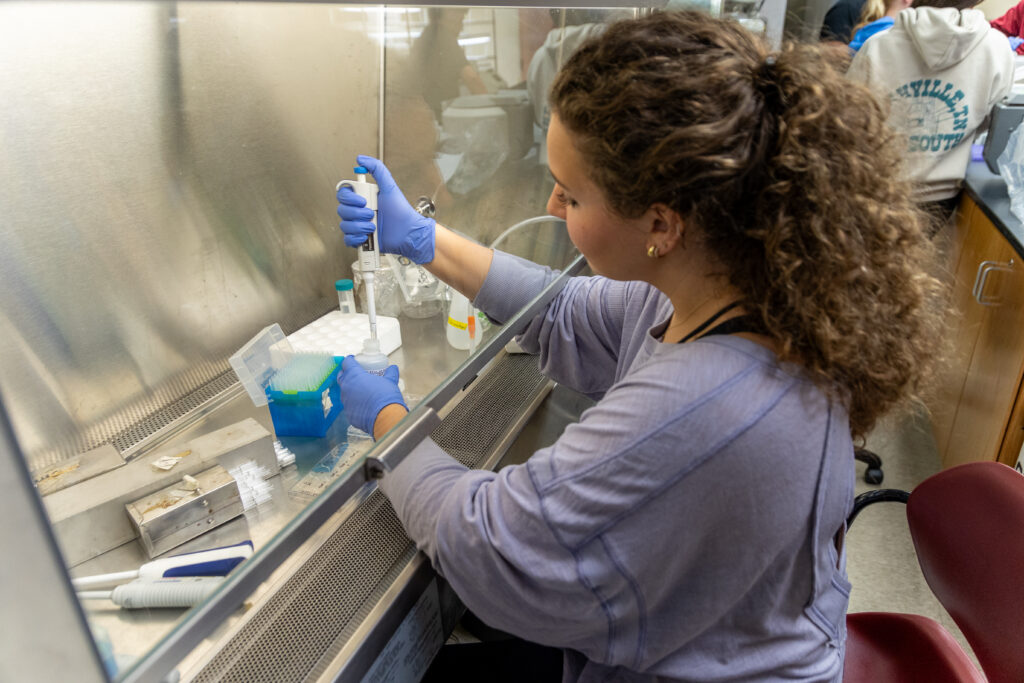A person with curly hair and blue gloves uses a pipette at a lab bench inside a fume hood, surrounded by cell biology equipment and supplies, with another person visible in the background.
