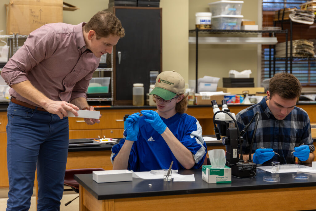 A teacher stands next to two students at a lab table, guiding them through a Conservation Biology experiment. Both students wear gloves and concentrate on analyzing objects, surrounded by lab equipment essential for studying biodiversity.