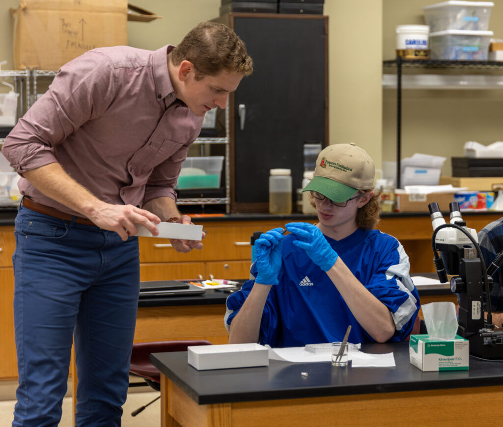 A student wearing gloves examines a small object at a lab table while an instructor stands nearby, holding a box and looking at the student. Lab equipment and supplies are visible in the background.