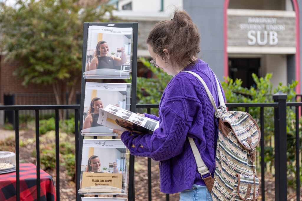 A person wearing a purple sweater and striped backpack stands outdoors reading a magazine next to a display rack, capturing a candid moment of photojournalism. A campus building is visible in the background.