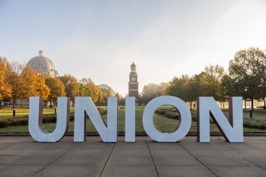 Large white letters spelling UNION stand on a pathway at Union's main campus in Jackson, with a domed building on the left and a clock tower in the center background, surrounded by Tennessee’s autumn foliage.