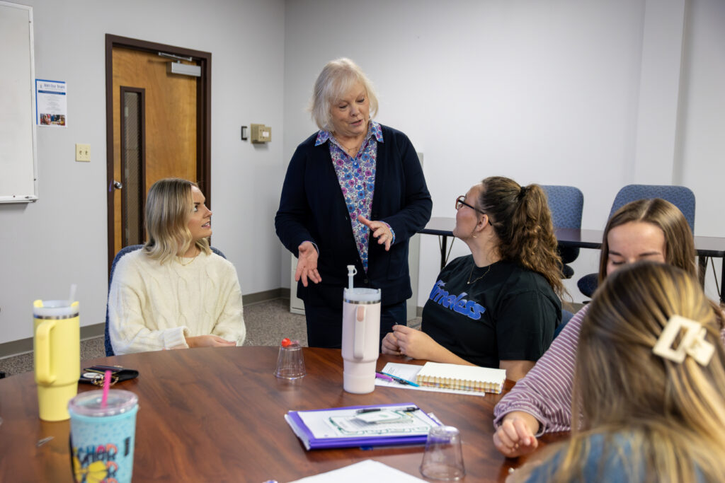 A woman stands and speaks to four seated women around a table in a classroom, likely discussing topics related to an MA in Education. The table holds notebooks, water bottles, and plastic cups as the group engages in discussion.