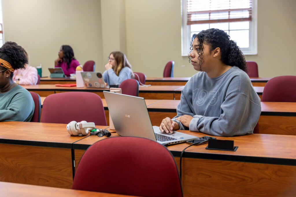 Students sit spaced out in a classroom with red chairs and wooden desks. One student in the foreground uses a laptop, while others are listening or working on their own devices. Sunlight enters through a window.