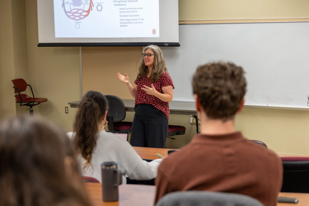 A woman stands at the front of a classroom giving an education-focused presentation to seated students. A projection screen behind her displays a diagram with text. Desks, chairs, and a whiteboard are also visible in the room.