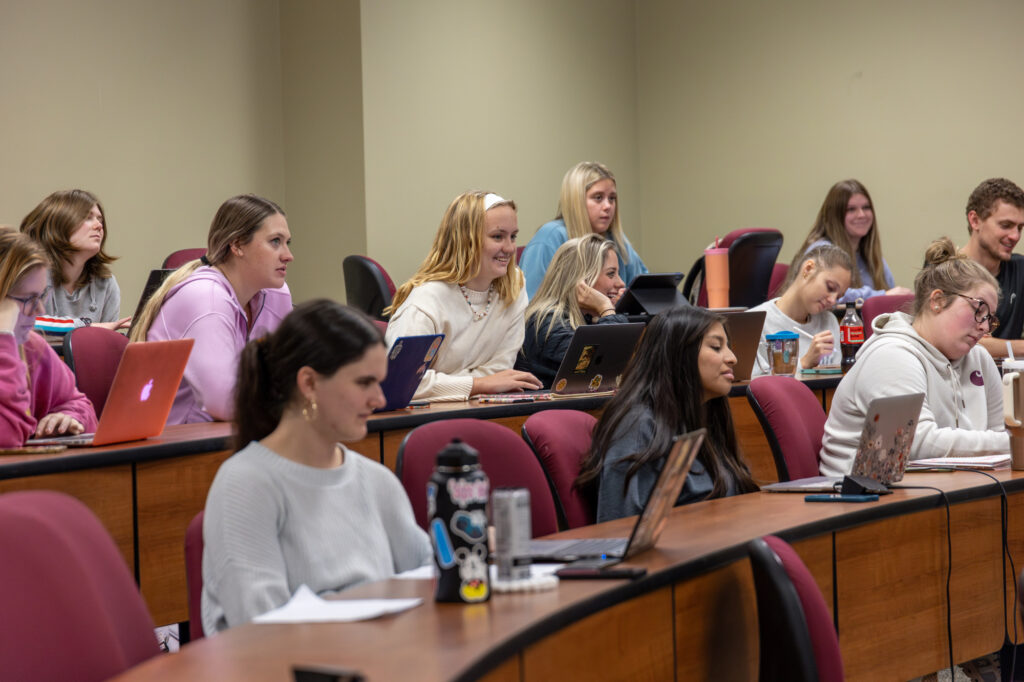 Students sit in a classroom with laptops open, seated at curved desks. Some are taking notes while others are listening. The room has red chairs and beige walls. The mood appears focused and engaged.