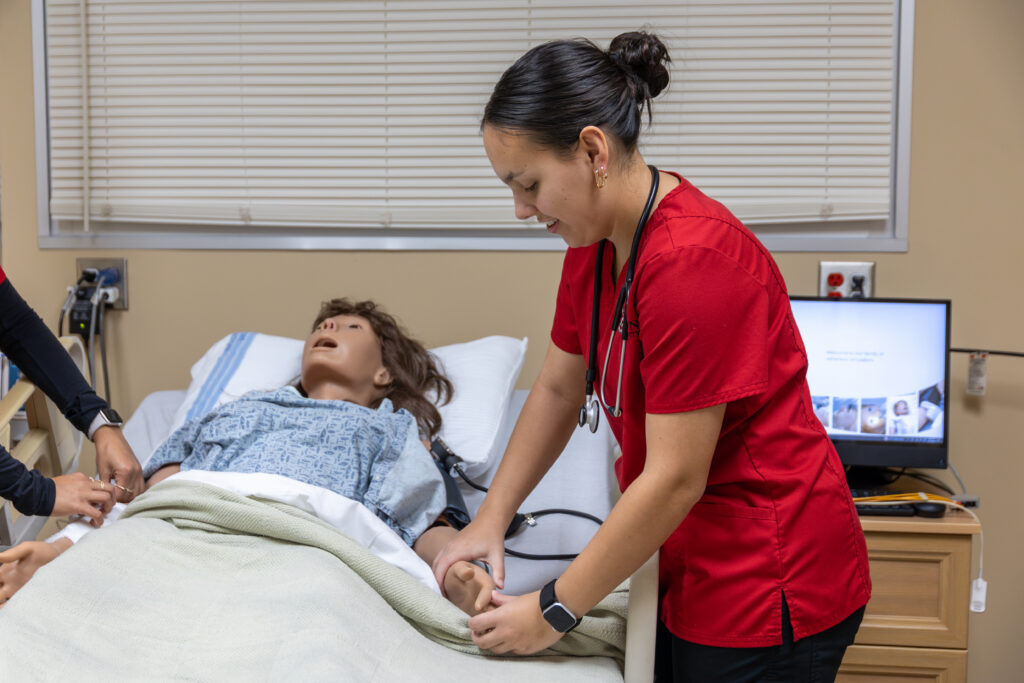A person in a red scrub top, possibly an accelerated BSN student, stands beside a medical mannequin lying in a hospital bed, adjusting the blanket. Medical equipment and a computer are visible in the background.
