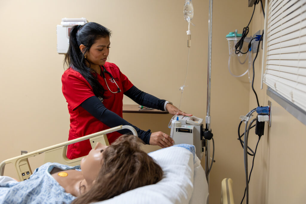 A DNP student specializing in Nurse Anesthesia, dressed in red scrubs, adjusts an IV machine beside a hospital bed where a medical mannequin lies under a blanket, simulating a patient in a clinical training room.