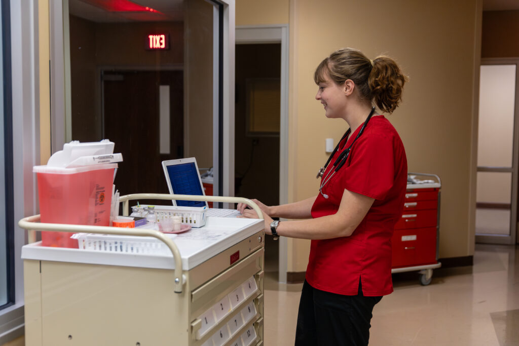 A nurse in red scrubs, exemplifying executive leadership, stands at a medical cart, looking at a laptop screen. The cart holds medical supplies and containers. She is in a hallway with beige walls and an open door in the background.
