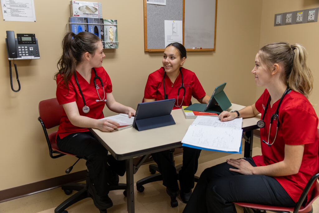 Three people in red scrubs and stethoscopes sit around a table with tablets and papers, discussing Health Management strategies in a small room with beige walls and medical equipment.
