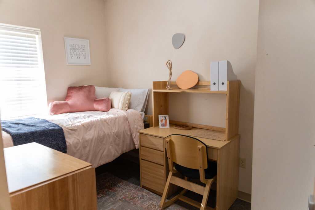 A small bedroom with a neatly made bed, pink pillows, a wooden desk with a chair, two white binders, a small framed photo, and a window with blinds letting in natural light.