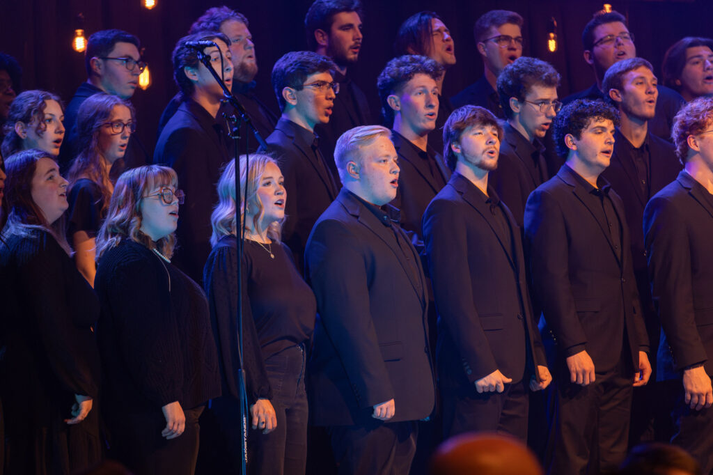 A choir of men and women dressed in black deliver a captivating performance on stage in front of microphones, with warm stage lights and hanging bulbs illuminating the background.