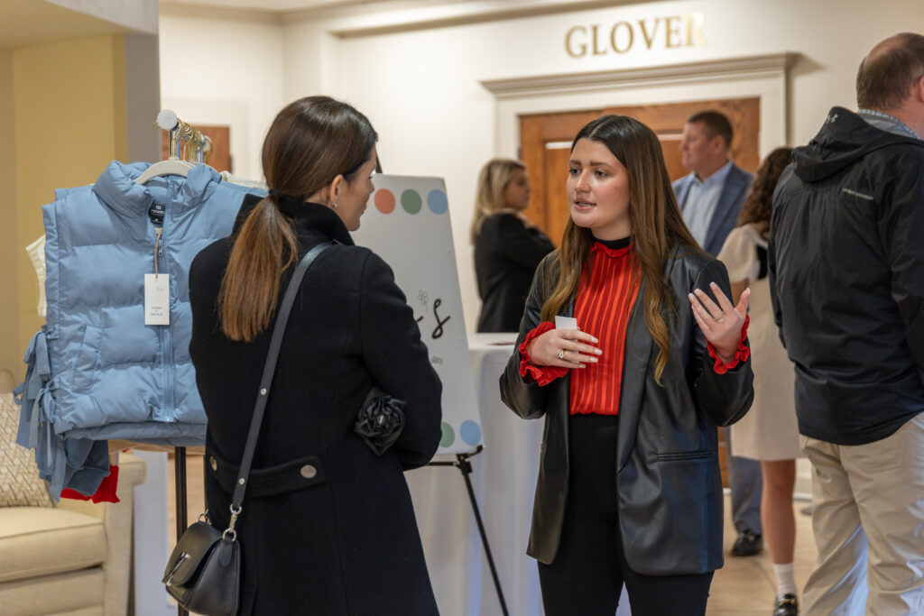 Two women, possibly discussing Business Administration or Marketing, stand and talk in a room with others. One wears a red blouse and black blazer; the other, a dark coat. A clothing rack with a blue vest and a sign are nearby. 