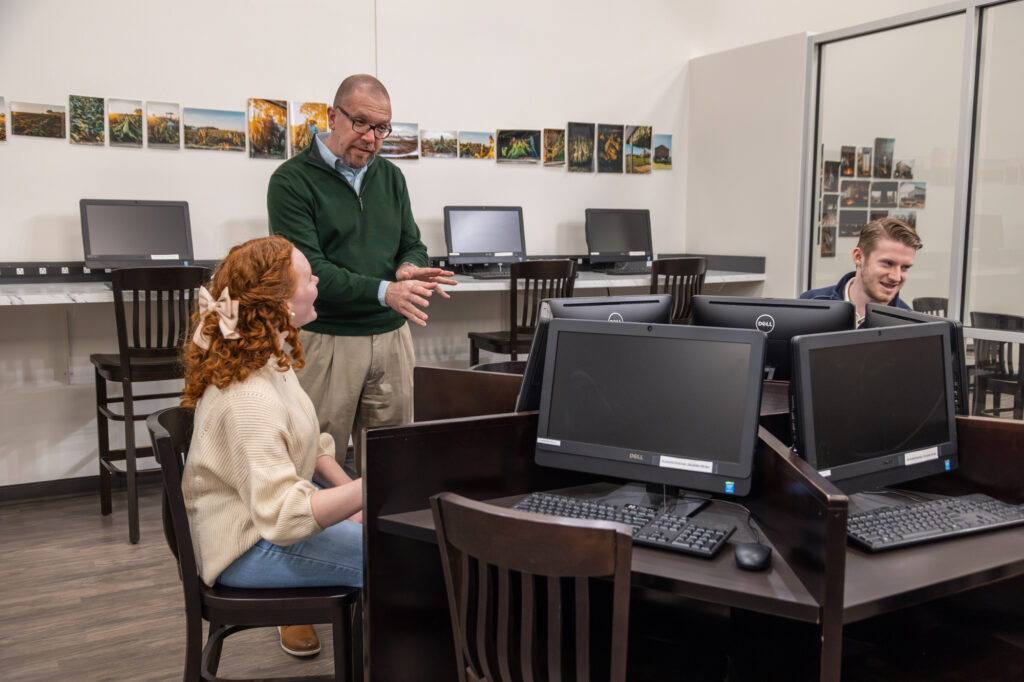 A man stands and speaks to a seated woman and man in a computer lab during an Honors Integrative Studies session. The woman faces him while the other man looks at a computer. Several landscape photos are displayed on the wall behind them.