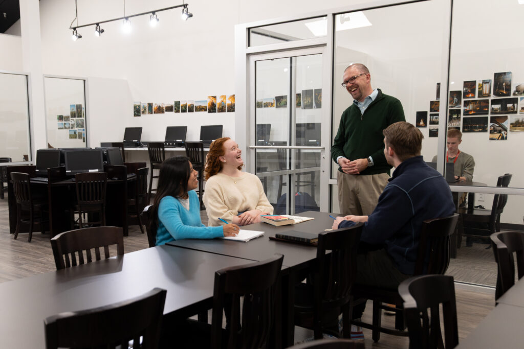 Four people sit at a table in a classroom, talking and smiling. One person is standing and engaging with the group. The room has computers, chairs, and photos on the wall. Another person works alone in the background.