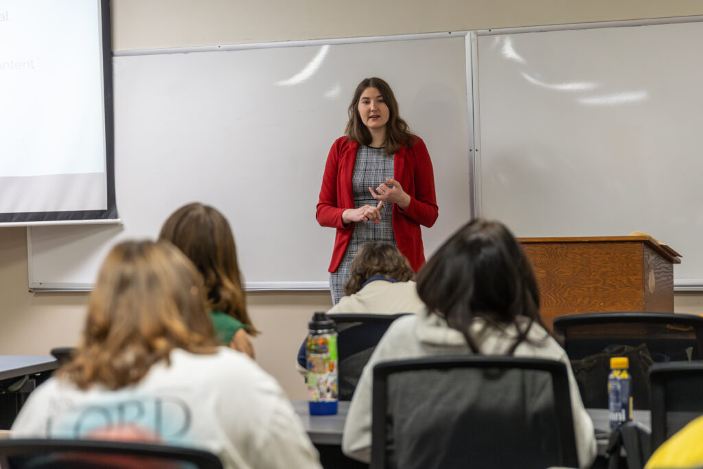 A woman in a red blazer stands at the front of a Family Studies classroom, speaking to students who are seated and facing her. A whiteboard and a wooden podium are visible in the background.