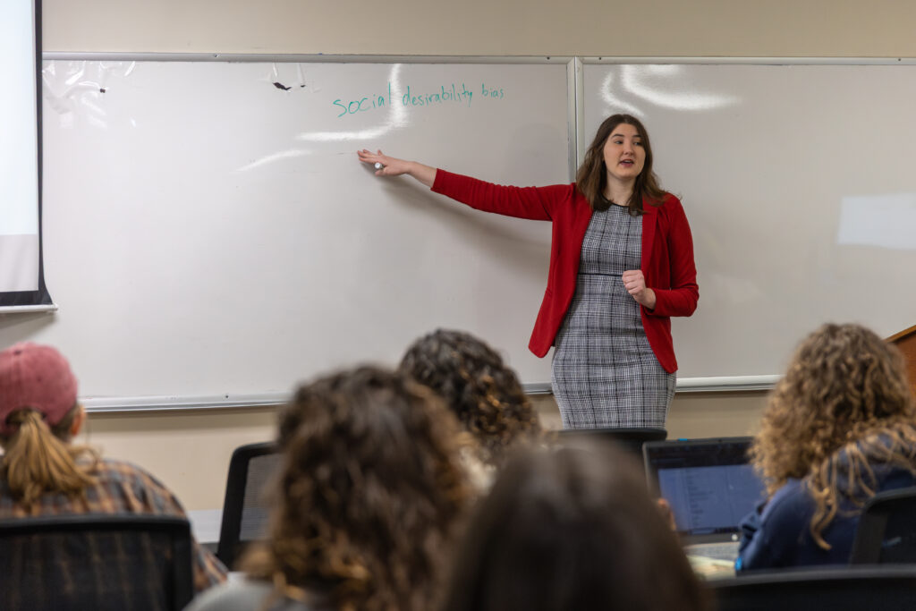 A woman in a red blazer stands at the front of a Family Studies classroom, pointing to a whiteboard with text partially reading Social desirability bias, while students sit facing her.
