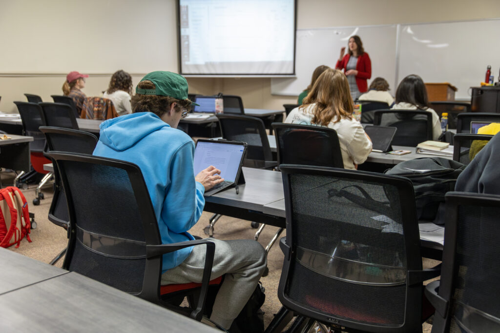 A classroom with students seated at desks, some using laptops and tablets. A teacher stands at the front near a whiteboard and a projection screen, speaking to the class about topics in Family Studies and Pre-Counseling.