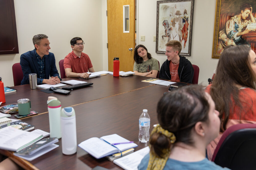 Six people sit around a conference table with notebooks, water bottles, and papers in front of them, engaged in discussion. The group appears to be meeting for a Renaissance Studies seminar. Two paintings are on the wall and a closed door is in the background.