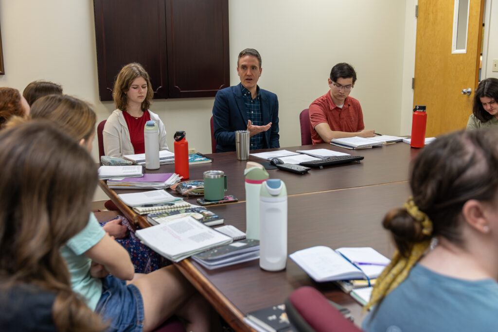 A group of people sit around a conference table with notebooks, water bottles, and papers while a man in a blue blazer speaks. Most attendees are listening, suggesting an English or literature-focused meeting or classroom setting.