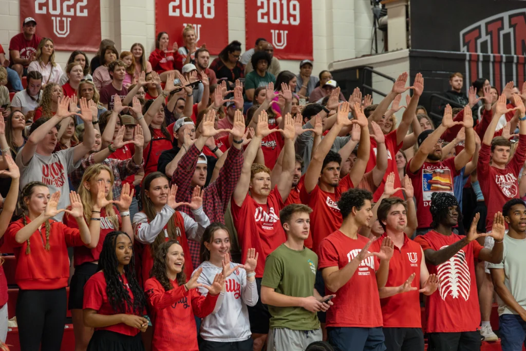 A large group of students in red and white shirts stand in bleachers, many holding their hands up in a U shape. Banners with years from 2017 to 2022 hang on the wall behind them.