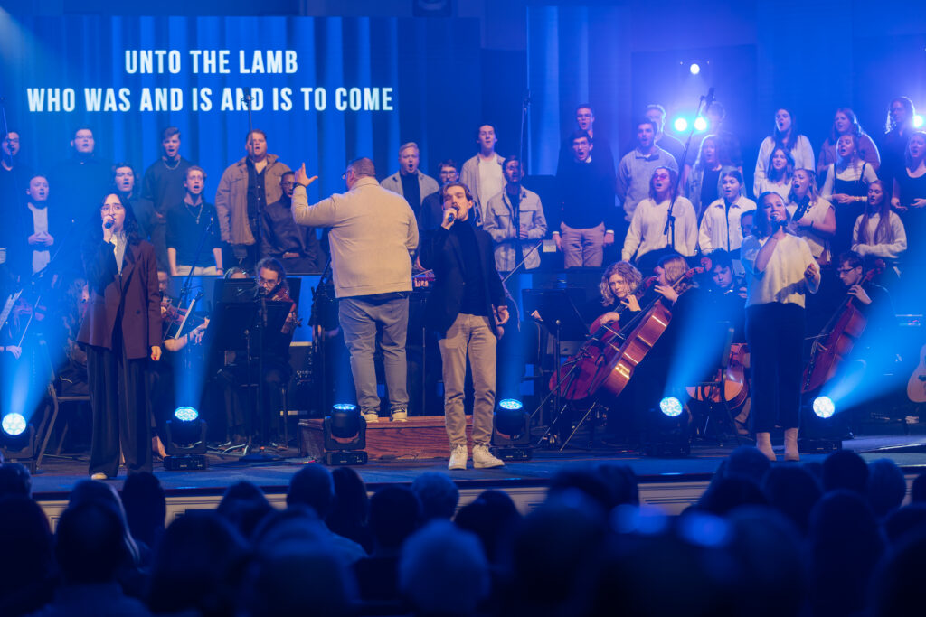 A choir and orchestra perform on stage in front of an audience, with four vocalists at the front. A screen behind them displays the words, “UNTO THE LAMB WHO WAS AND IS AND IS TO COME.” Blue stage lighting is used.