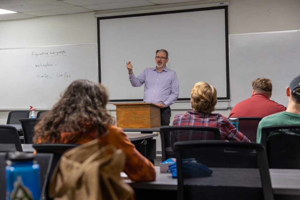 A man stands at a podium in front of a classroom, speaking to students about applied linguistics. Writing covers the whiteboard behind him, and a projector screen hangs above as the students face the front attentively.