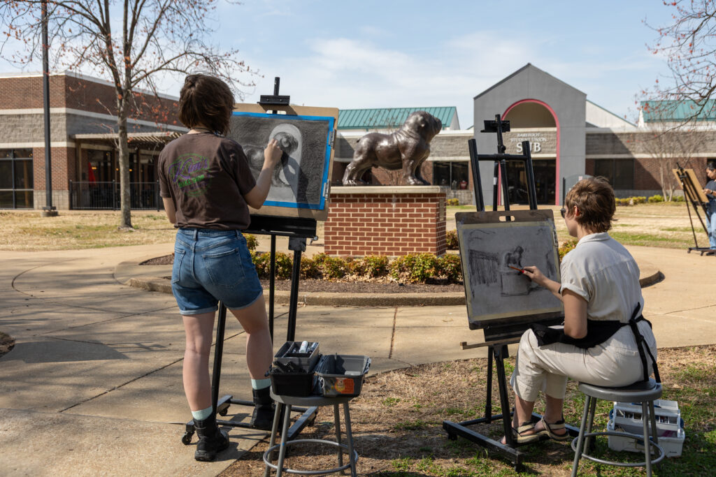 Two people enjoy drawing at easels outdoors in front of a building with a dog statue. One stands while sketching, the other sits on a stool, surrounded by art supplies scattered on the ground.