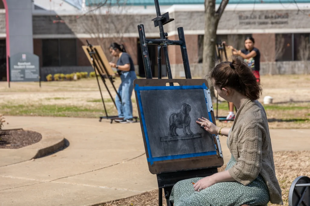 Several people draw at easels outdoors. In the foreground, a woman sketches a black and white image of a gorilla on paper attached to an easel. Other artists work in the background near a building.