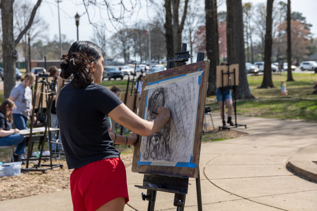A person wearing a black shirt and red shorts creates art on a large sketchpad mounted on an easel outdoors, surrounded by others drawing in a park-like setting.