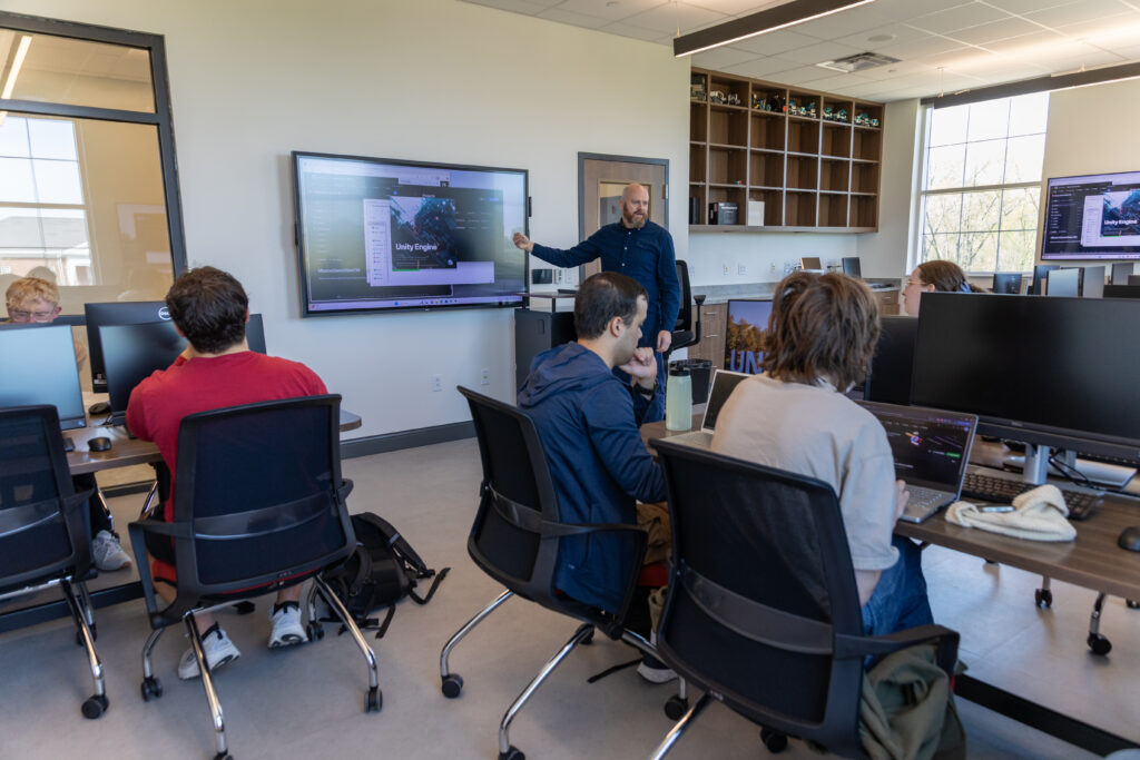 A man stands at a screen displaying a computer science presentation at the front of a classroom while several students seated at desks with computers listen and work.