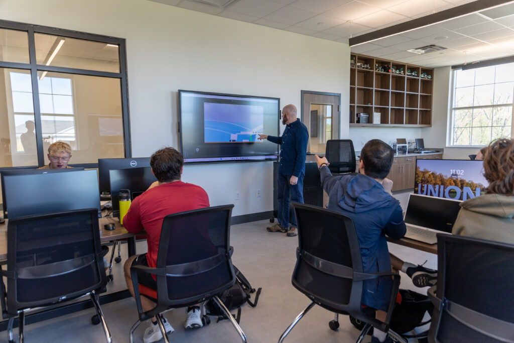 A person stands at a large screen displaying a technical diagram related to computer science, pointing at it, while several people seated at desks with monitors watch and listen in a modern classroom setting.