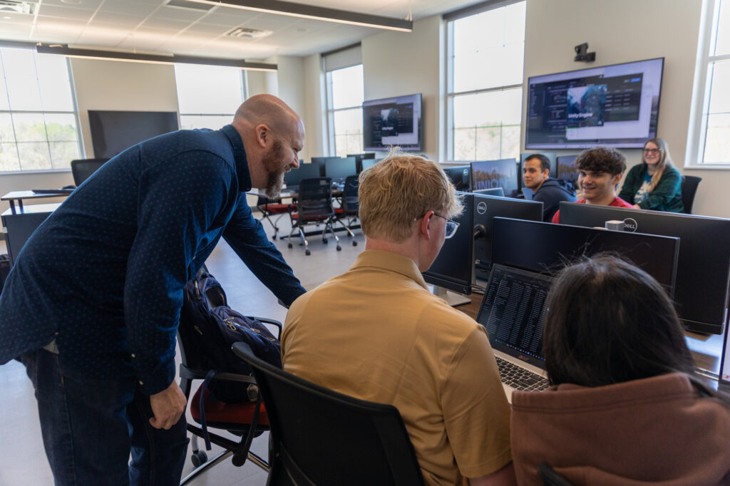 A teacher leans over a student’s desk in a computer science lab while other students sit at their computers, some looking at screens and some at the teacher. Large windows and monitors are visible in the background.