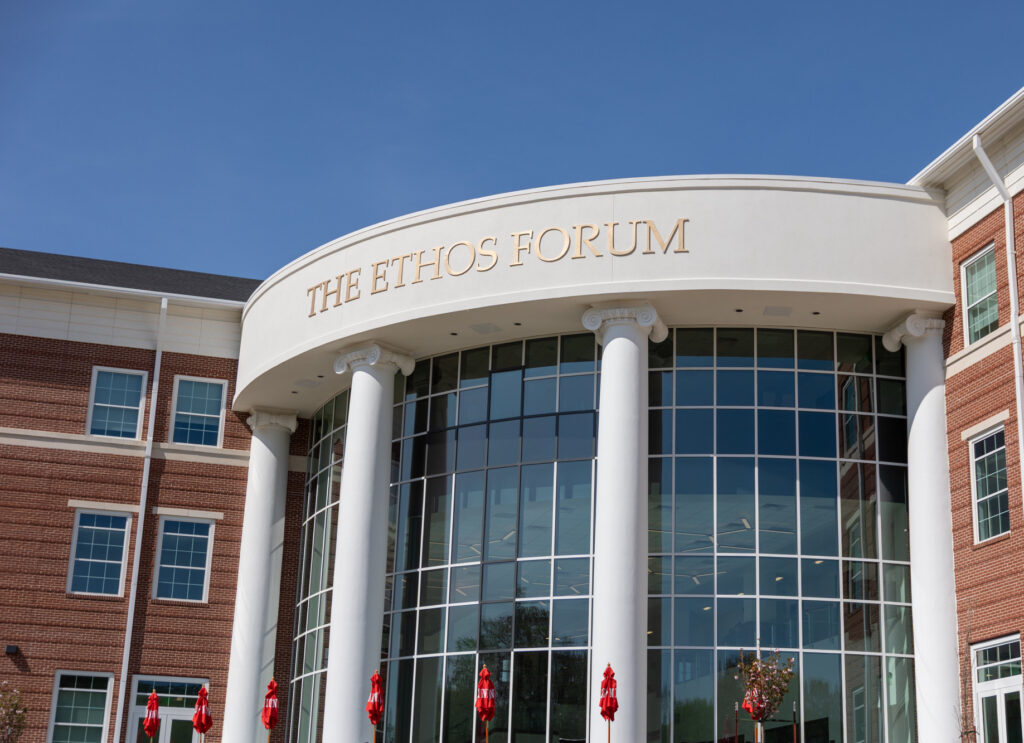 A modern brick building with large white columns and glass windows houses the McAfee School of Business. “THE ETHOS FORUM” is displayed in gold letters above the entrance, with red umbrellas visible outside.