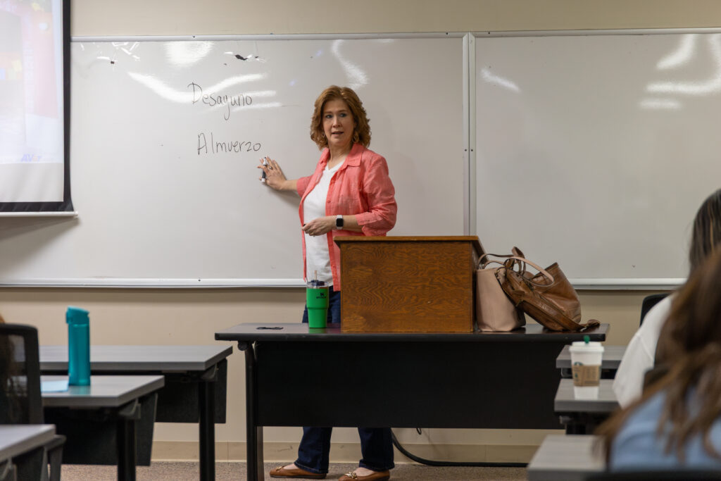 A woman stands at a whiteboard in a classroom, writing the words “Desayuno” and “Almuerzo.” She faces the students, with a wooden podium and bags beside her. Several desks, bottles, and students are visible in the foreground.