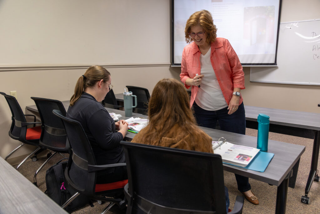 A woman stands and talks to two seated students in a French Studies classroom. The students have open notebooks and materials on their desks, with a whiteboard and projection screen visible in the background.