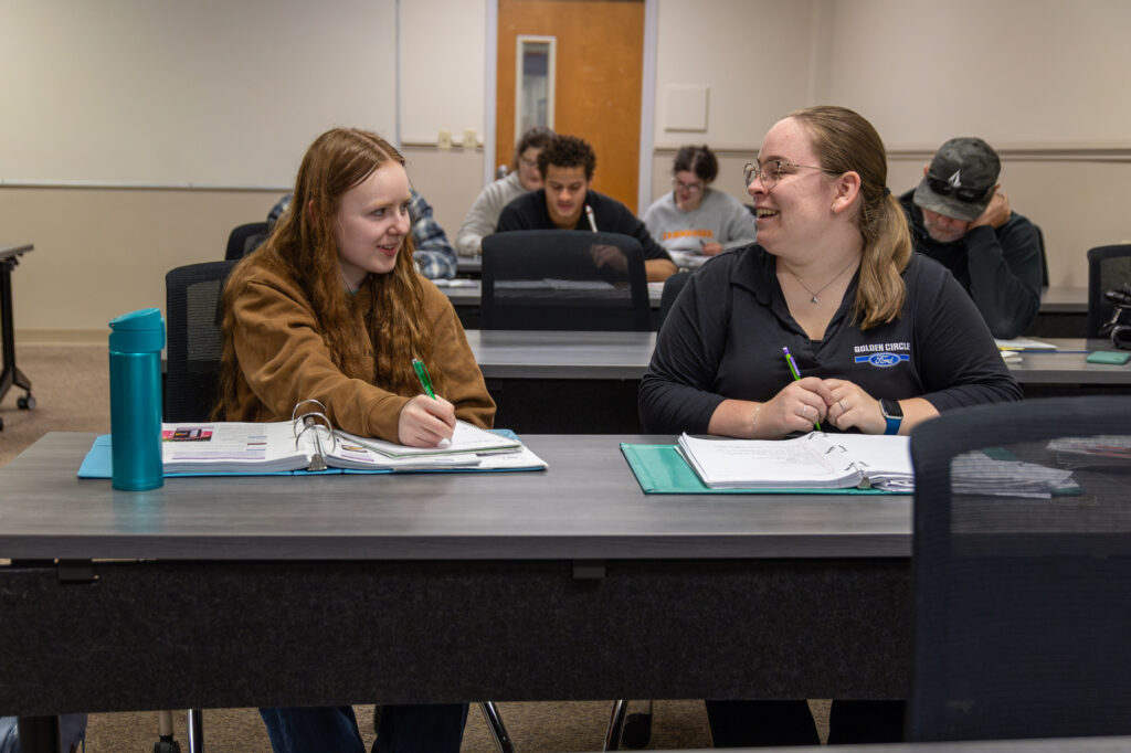 Two people sit at a classroom desk, writing in notebooks and smiling at each other. Other students are seated at desks in the background. A blue water bottle is on the desk in the foreground.
