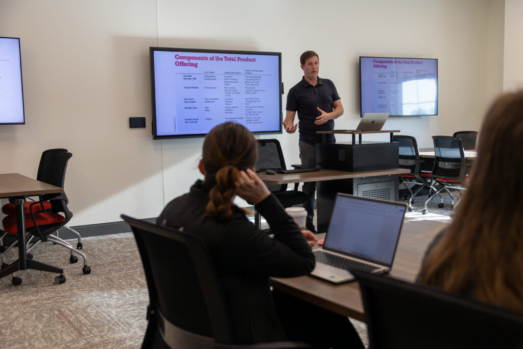 A man presents digital marketing insights on a screen to a group in a conference room. Attendees, possibly MBA students, sit at tables with laptops, facing the presenter. The room has neutral walls and multiple monitors displaying slides.