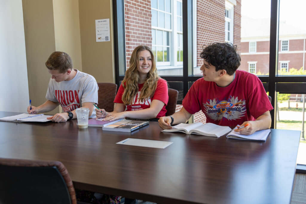 Three students sit at a table by large windows on Union's main campus in Jackson, Tennessee, studying and writing in notebooks. Two wear red shirts and one a white shirt. Books, papers, and drinks are on the table in the naturally lit, brick-walled room.