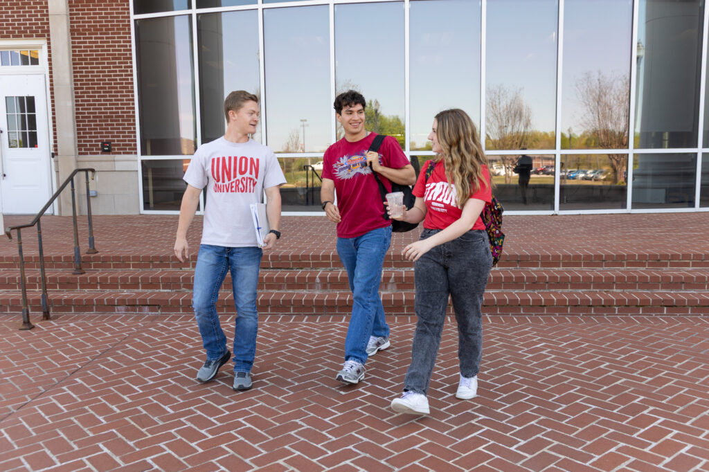Three students walk and talk together on a brick walkway in front of a modern building with large windows. Two students wear red university T-shirts, and one carries a notebook.
