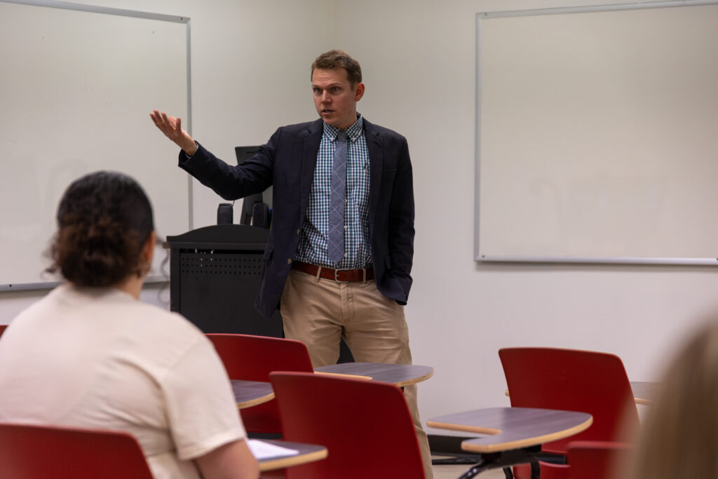 A man in a blazer and checked shirt stands at the front of a classroom, speaking to seated students. The classroom has red chairs, whiteboards, and a podium.