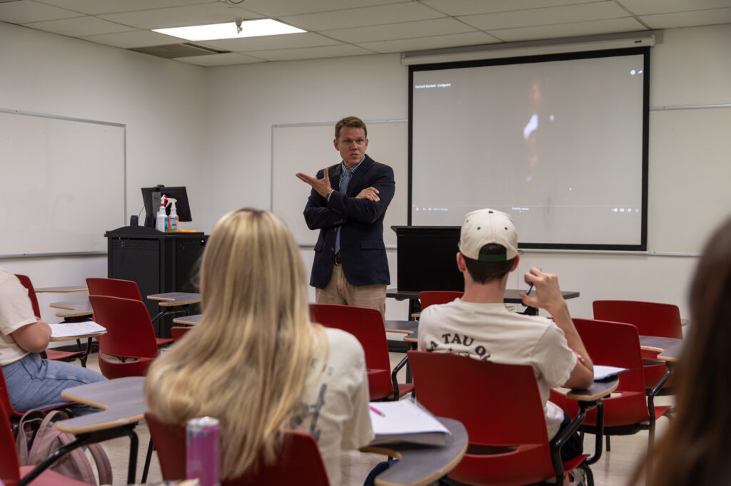 A man in a suit stands at the front of a classroom with red chairs, gesturing as he lectures on Applied Linguistics. A projector displays an image on the screen behind him while students face him attentively and take notes.