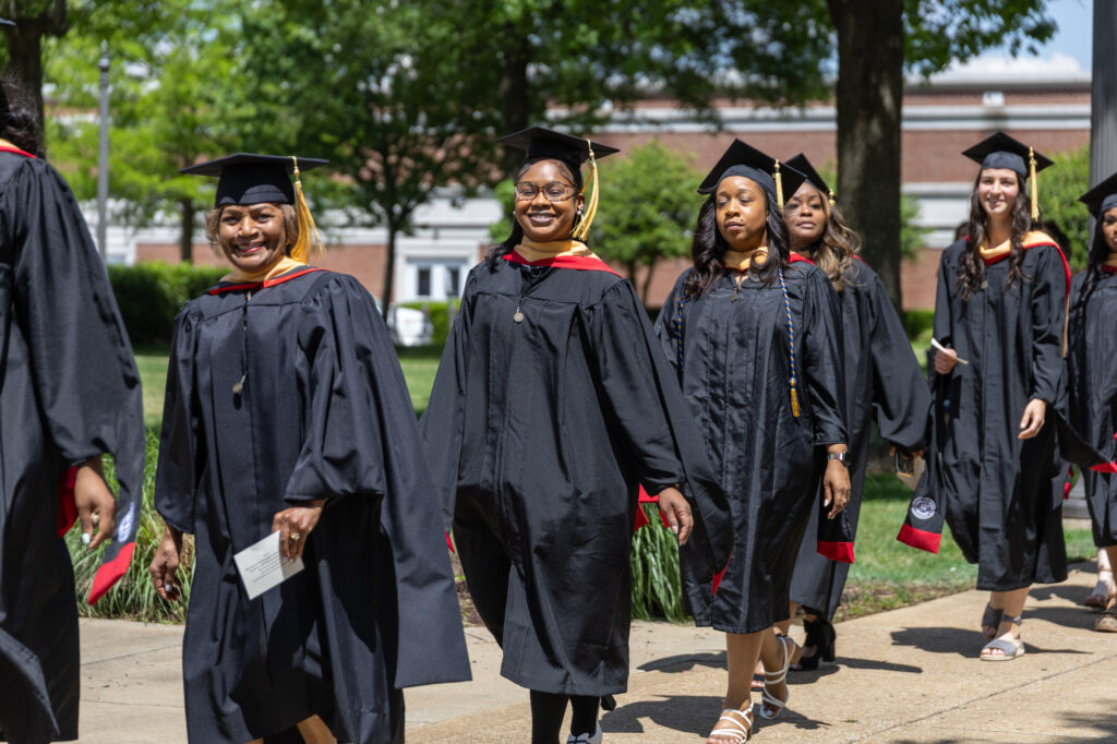 A group of graduates in black caps and gowns walk outdoors on a sunny day, smiling and heading in the same direction. Trees, grass, and a building are visible in the background.