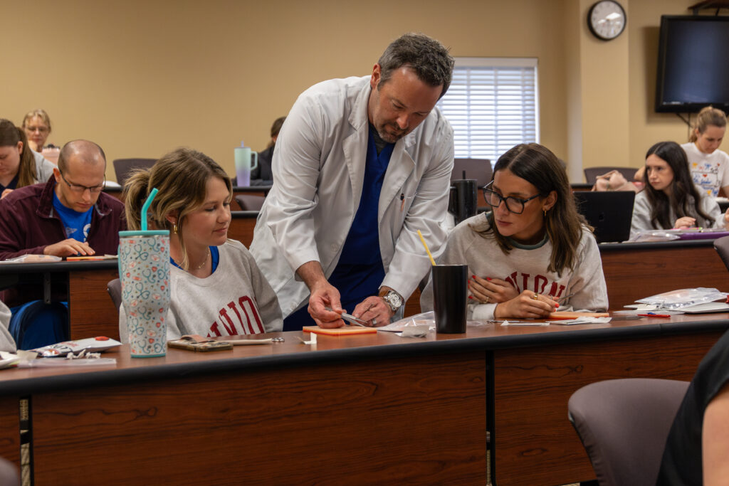 A man in a white lab coat, possibly a Doctor of Nursing Practice (DNP), instructs two women at a classroom desk while other students work in the background. The desks have supplies, drinks, and paperwork on them.