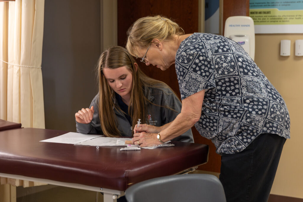 An older woman, likely a Nurse Practitioner, stands beside a younger woman seated at a table, both looking at papers. The older woman points to something on the paper amid medical supplies, suggesting an NPGC clinical setting.
