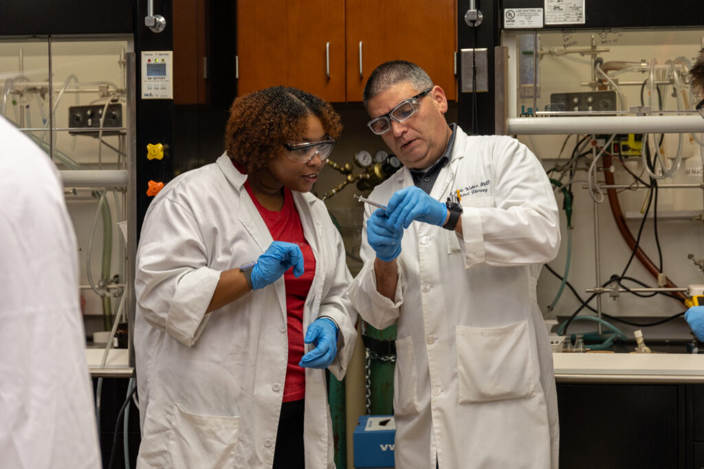 Two people with a PharmD, wearing lab coats, safety glasses, and blue gloves, examine a small object in a laboratory setting filled with scientific equipment and cabinets.