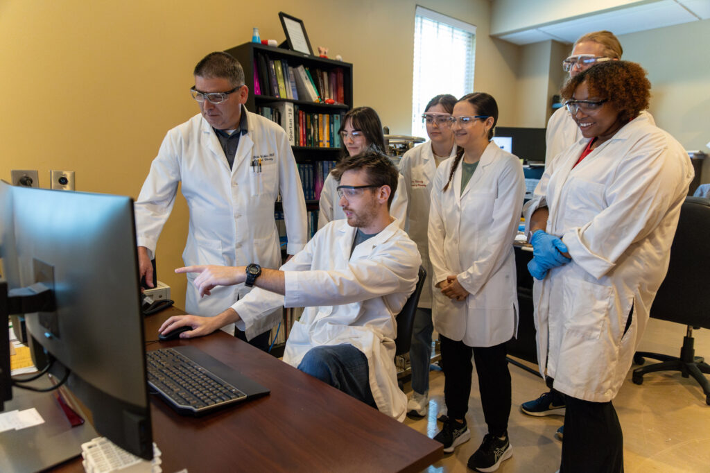 Six people in lab coats and safety glasses gather around a computer in an office, with one PharmD student seated and pointing at the monitor while the others stand and watch attentively. Bookshelves and desks are visible in the background.