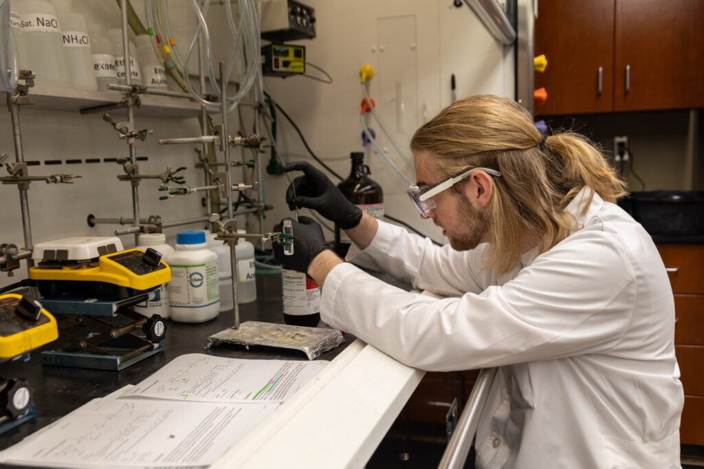 A person in a lab coat and safety goggles, holding a PharmD or pharmacy degree, works with lab equipment and chemicals on a laboratory bench, surrounded by documents, bottles, and scientific instruments.