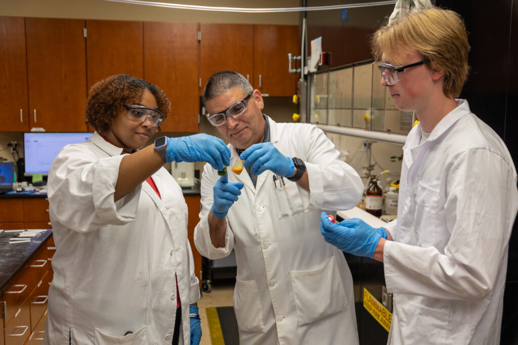 Three people with a pharmacy degree, wearing lab coats, safety glasses, and blue gloves, collaborate in a laboratory while examining a test tube with yellow liquid. Laboratory equipment and cabinets are visible in the background.