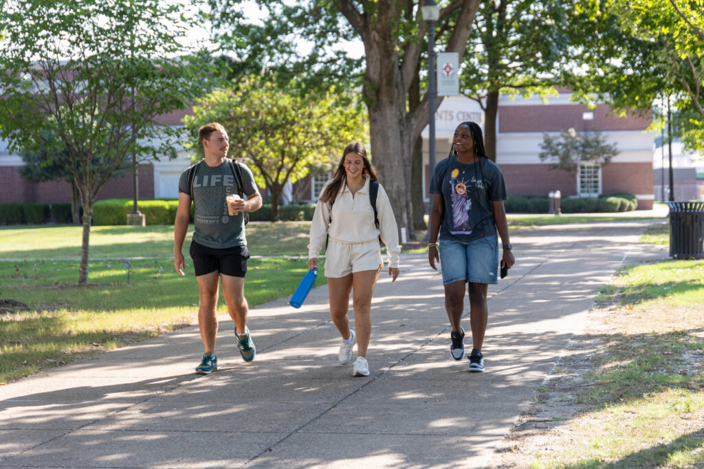 Three people walk together on a shaded campus pathway at Union's main campus in Jackson. One holds a drink, another carries a blue water bottle, and all are dressed in casual summer clothes. Trees and a brick building are visible in the background.